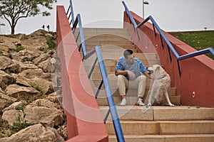 Young Hispanic man, sitting on stairs next to his dog while talking with him in a loving and tender attitude. Concept, dogs, pets