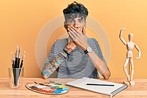 Young hispanic man sitting on the table at art studio shocked covering mouth with hands for mistake