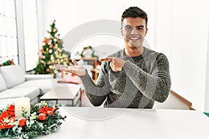 Young hispanic man sitting on the table by christmas tree amazed and smiling to the camera while presenting with hand and pointing