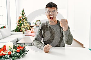 Young hispanic man sitting on the table by christmas tree angry and mad raising fist frustrated and furious while shouting with