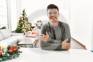 Young hispanic man sitting on the table by christmas tree happy face smiling with crossed arms looking at the camera