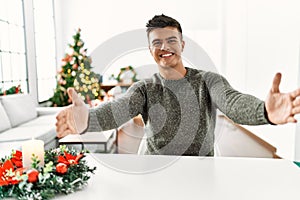 Young hispanic man sitting on the table by christmas tree looking at the camera smiling with open arms for hug