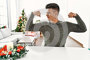 Young hispanic man sitting on the table by christmas tree showing arms muscles smiling proud