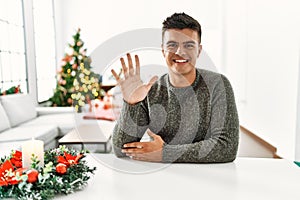 Young hispanic man sitting on the table by christmas tree showing and pointing up with fingers number five while smiling confident