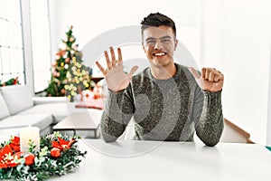 Young hispanic man sitting on the table by christmas tree showing and pointing up with fingers number six while smiling confident