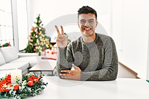 Young hispanic man sitting on the table by christmas tree showing and pointing up with fingers number two while smiling confident