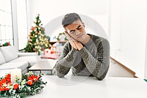 Young hispanic man sitting on the table by christmas tree sleeping tired dreaming and posing with hands together while smiling