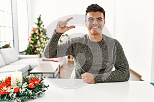 Young hispanic man sitting on the table by christmas tree smiling and confident gesturing with hand doing small size sign with