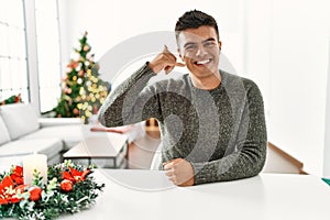 Young hispanic man sitting on the table by christmas tree smiling doing phone gesture with hand and fingers like talking on the