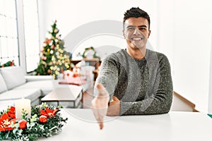 Young hispanic man sitting on the table by christmas tree smiling friendly offering handshake as greeting and welcoming