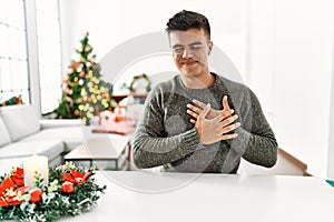 Young hispanic man sitting on the table by christmas tree smiling with hands on chest with closed eyes and grateful gesture on
