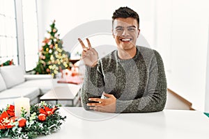 Young hispanic man sitting on the table by christmas tree smiling with happy face winking at the camera doing victory sign