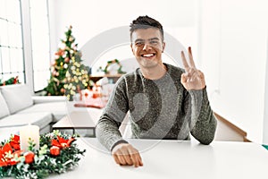 Young hispanic man sitting on the table by christmas tree smiling looking to the camera showing fingers doing victory sign