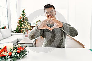 Young hispanic man sitting on the table by christmas tree smiling in love doing heart symbol shape with hands