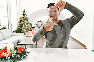 Young hispanic man sitting on the table by christmas tree smiling making frame with hands and fingers with happy face