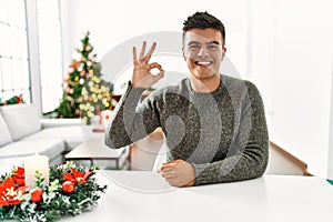 Young hispanic man sitting on the table by christmas tree smiling positive doing ok sign with hand and fingers