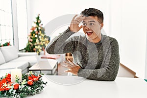 Young hispanic man sitting on the table by christmas tree very happy and smiling looking far away with hand over head