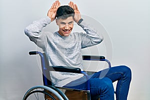 Young hispanic man sitting on wheelchair doing bunny ears gesture with hands palms looking cynical and skeptical