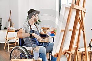 Young hispanic man sitting on wheelchair painting at art studio looking to side, relax profile pose with natural face and