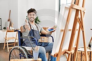 Young hispanic man sitting on wheelchair painting at art studio smiling cheerful offering palm hand giving assistance and