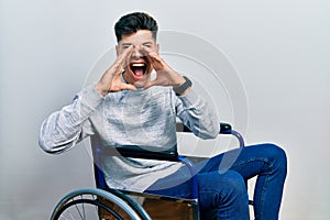 Young hispanic man sitting on wheelchair shouting angry out loud with hands over mouth