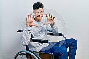 Young hispanic man sitting on wheelchair smiling funny doing claw gesture as cat, aggressive and sexy expression