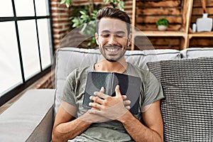 Young hispanic man smiling confident hugging bible at home