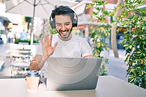 Young hispanic man smiling happy doing video call using laptop and headphones at coffee shop terrace