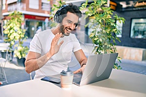 Young hispanic man smiling happy doing video call using laptop and headphones at coffee shop terrace