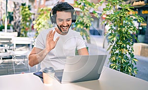 Young hispanic man smiling happy doing video call using laptop and headphones at coffee shop terrace