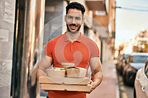 Young hispanic man smiling happy holding take away food at the city