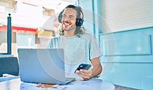 Young hispanic man smiling happy working using laptop and headphones at terrace of coffee shop