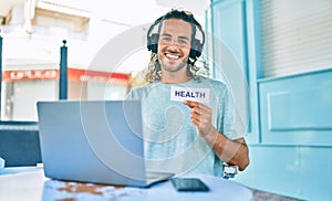 Young hispanic man smiling happy working using laptop and holding health word paper at terrace of coffee shop