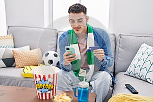 Young hispanic man soccer fan using smartphone and credit card sitting on sofa at home