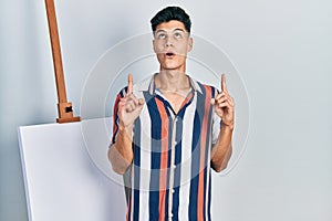 Young hispanic man standing close to empty canvas amazed and surprised looking up and pointing with fingers and raised arms