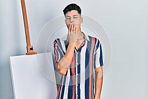 Young hispanic man standing close to empty canvas bored yawning tired covering mouth with hand