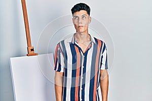 Young hispanic man standing close to empty canvas making fish face with lips, crazy and comical gesture