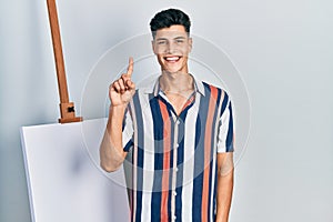 Young hispanic man standing close to empty canvas showing and pointing up with finger number one while smiling confident and happy