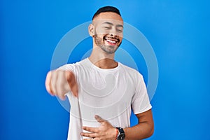 Young hispanic man standing over blue background laughing at you, pointing finger to the camera with hand over body, shame