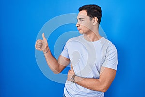 Young hispanic man standing over blue background looking proud, smiling doing thumbs up gesture to the side