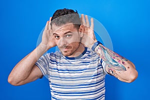 Young hispanic man standing over blue background trying to hear both hands on ear gesture, curious for gossip