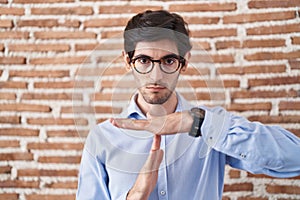 Young hispanic man standing over brick wall background doing time out gesture with hands, frustrated and serious face
