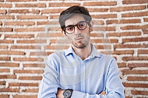 Young hispanic man standing over brick wall background skeptic and nervous, disapproving expression on face with crossed arms