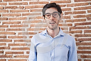 Young hispanic man standing over brick wall background winking looking at the camera with sexy expression, cheerful and happy face