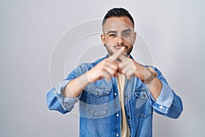 Young hispanic man standing over isolated background rejection expression crossing fingers doing negative sign