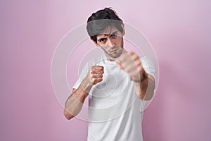 Young hispanic man standing over pink background punching fist to fight, aggressive and angry attack, threat and violence