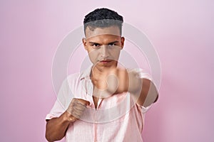 Young hispanic man standing over pink background punching fist to fight, aggressive and angry attack, threat and violence