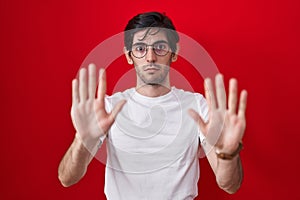 Young hispanic man standing over red background moving away hands palms showing refusal and denial with afraid and disgusting