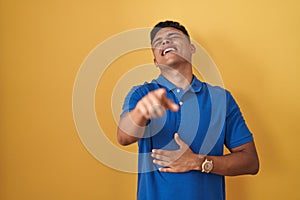 Young hispanic man standing over yellow background laughing at you, pointing finger to the camera with hand over body, shame