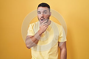 Young hispanic man standing over yellow background looking fascinated with disbelief, surprise and amazed expression with hands on
